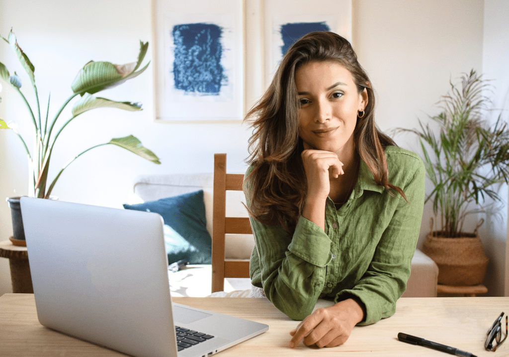 Person with long, light brown hair, wearing a green button down, sitting in front of a lap top, with art and plants in the background.